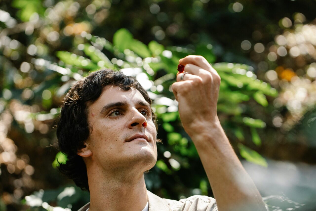 A contemplative man observes a small red berry under sunlight in a lush forest setting.